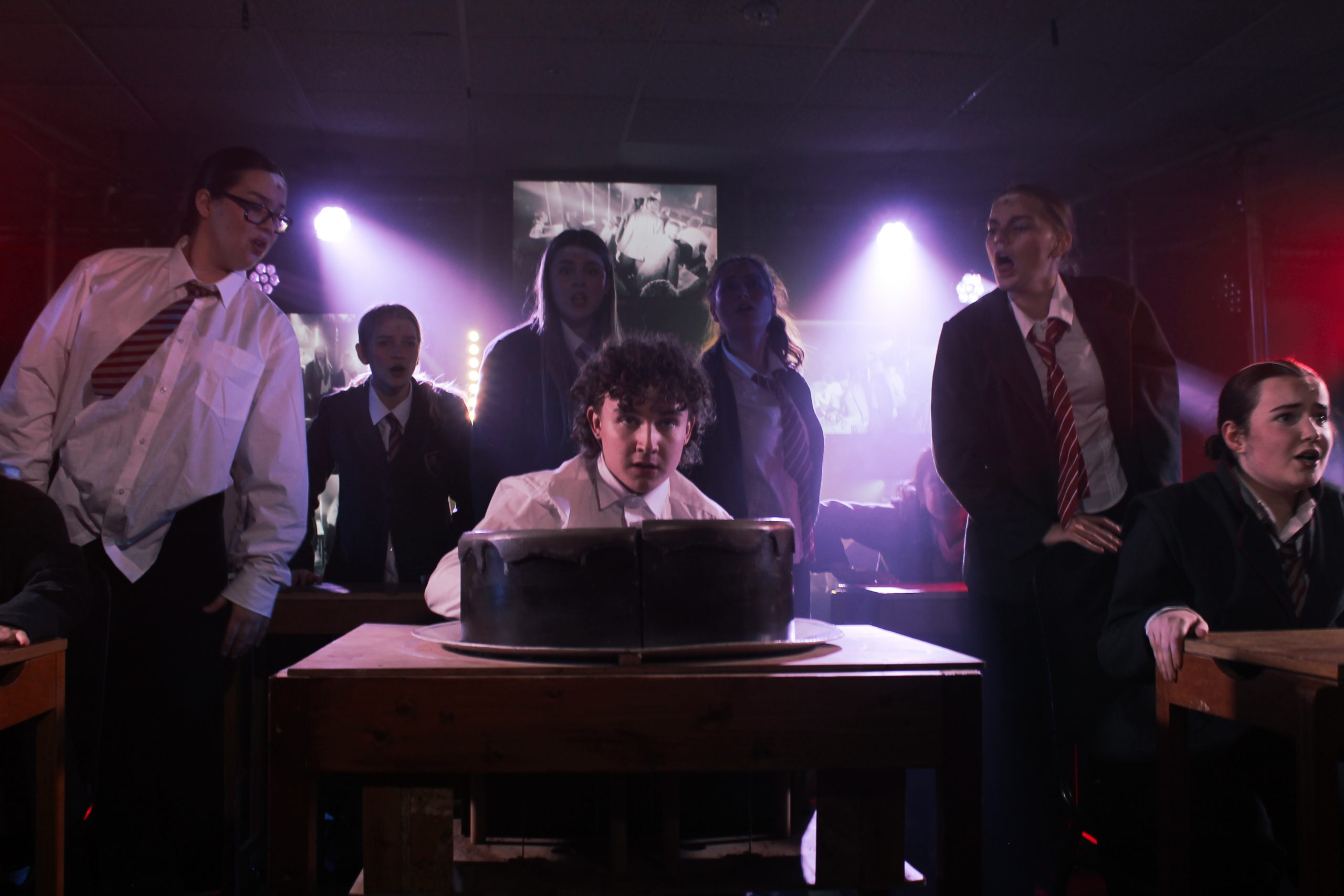 Pupils in school uniform gather around a boy seated behind a giant chocolate cake on a desk.