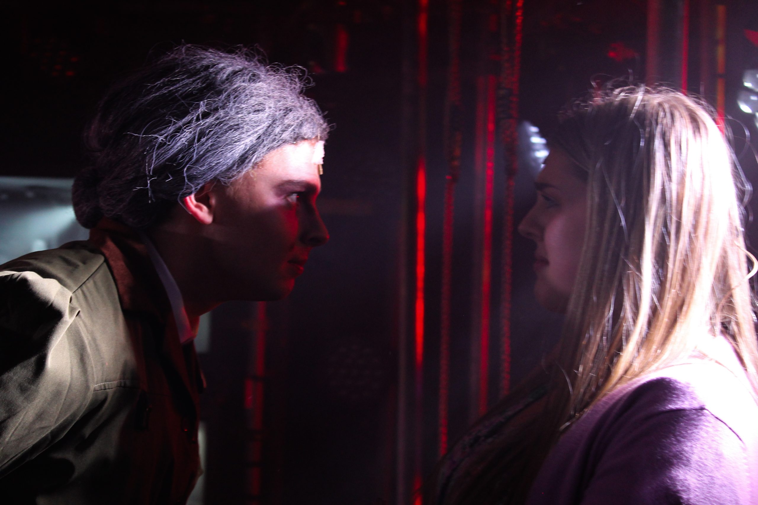 A performer in a grey wig confronts a pupil face to face in red light.