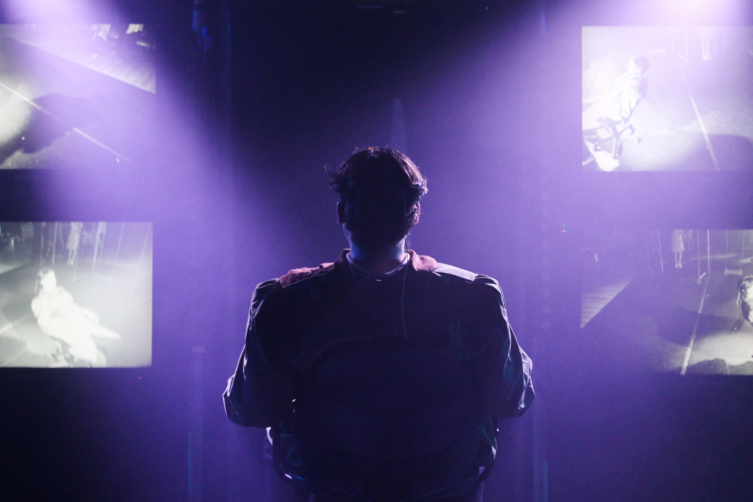 A figure seen from behind sits facing projected black-and-white images in purple haze.