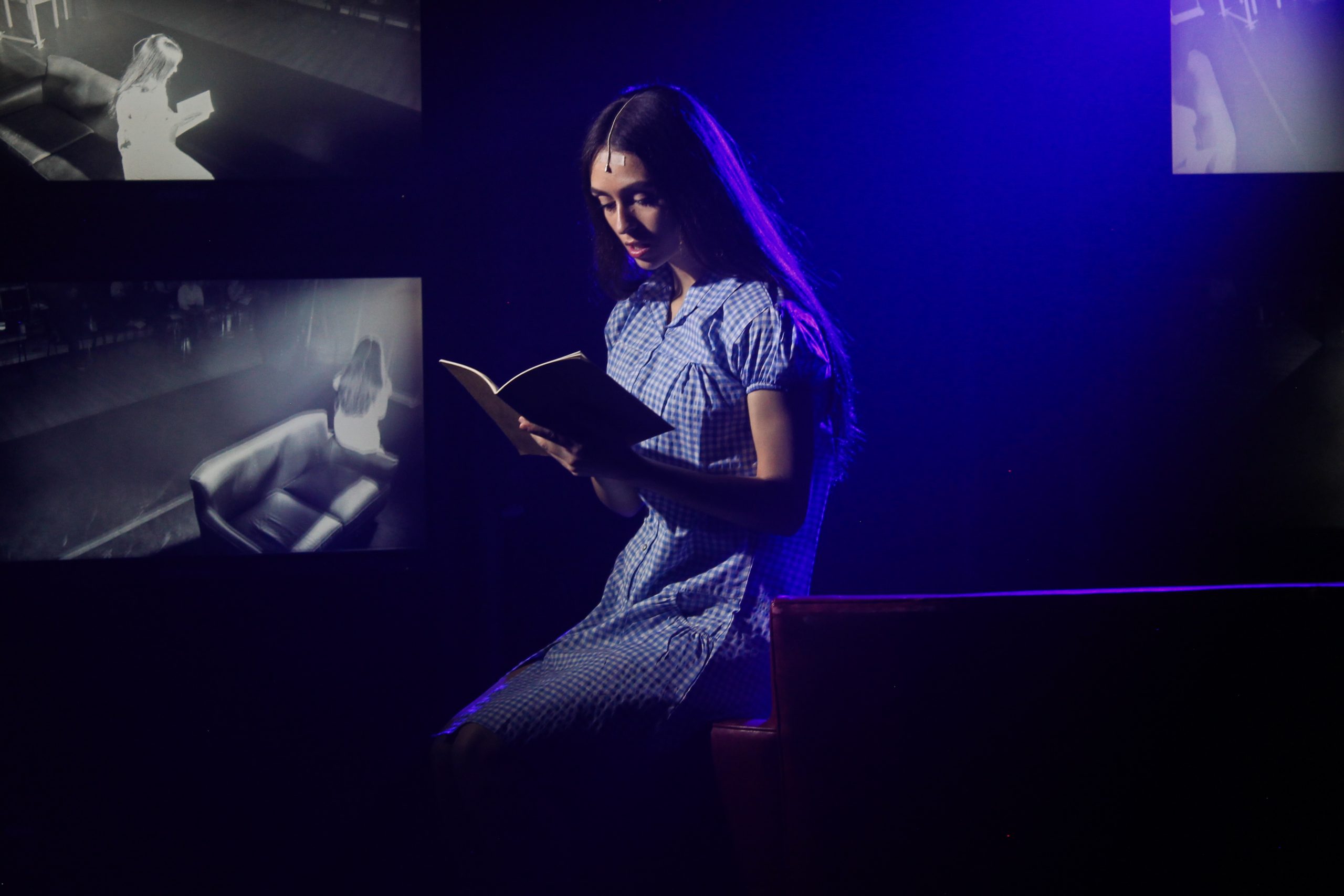 A girl in a blue gingham dress reads a book, with black-and-white images projected behind her.