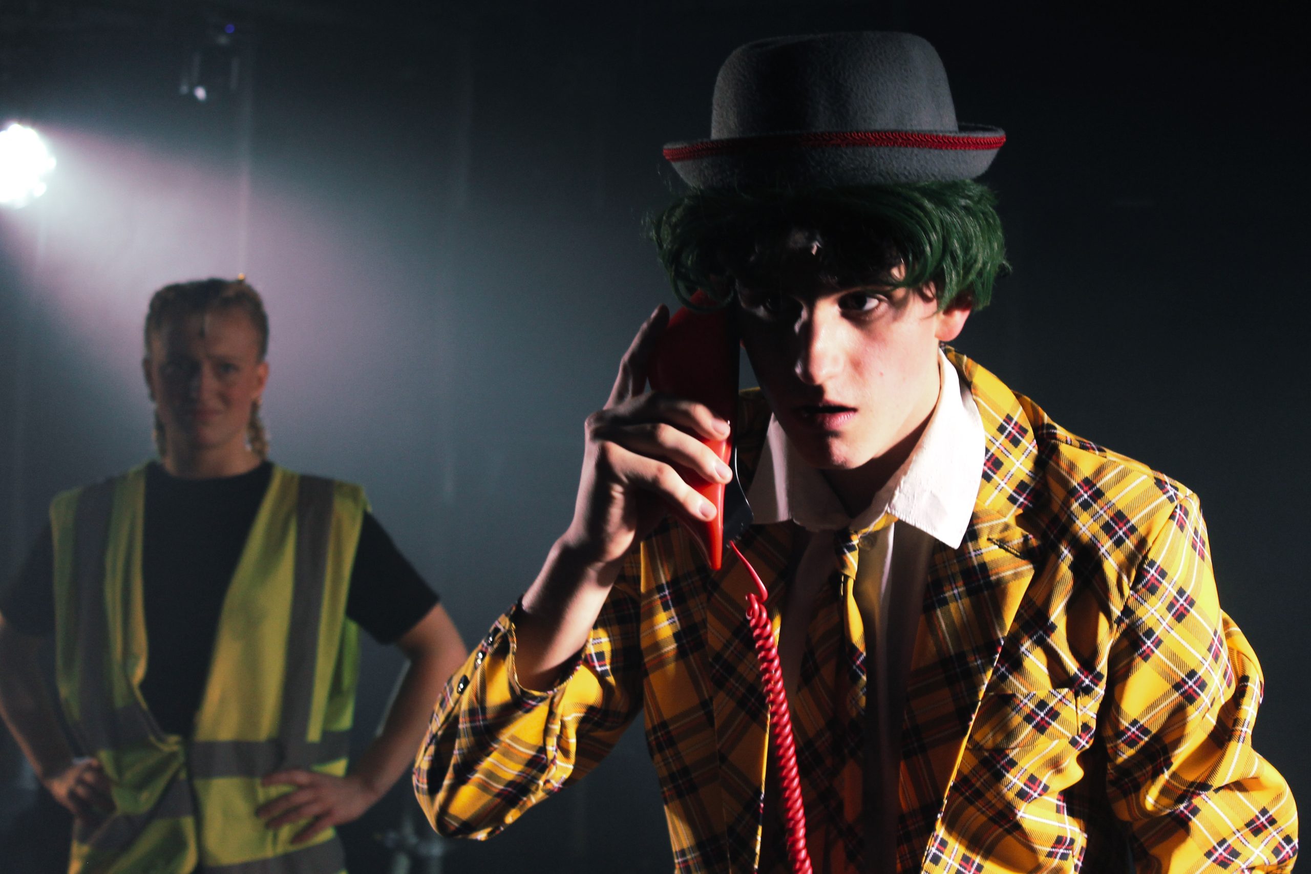 A performer in a yellow checked jacket and bowler hat talks on a red telephone, with a performer in a hi-vis vest behind.