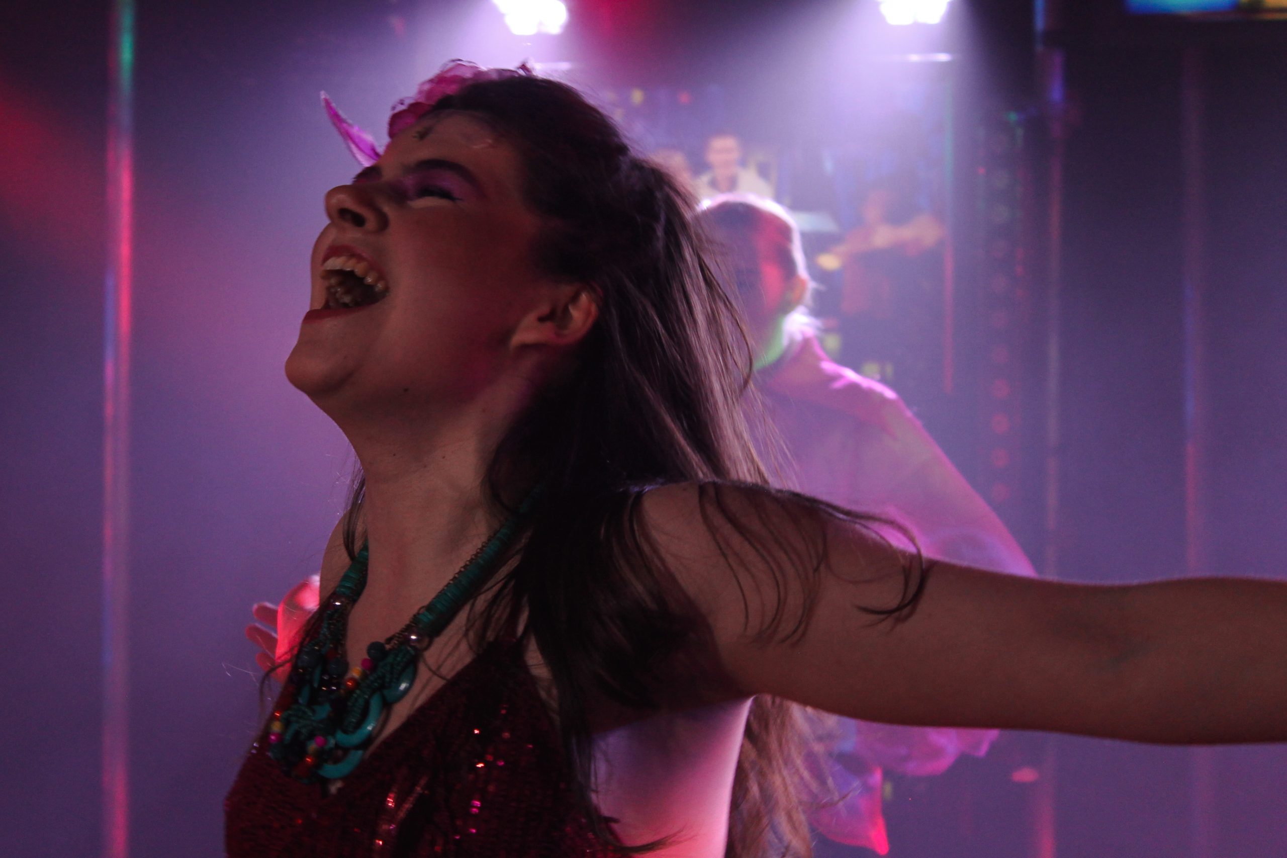 A performer in a sequinned top and beaded necklace throws her head back laughing in pink light.