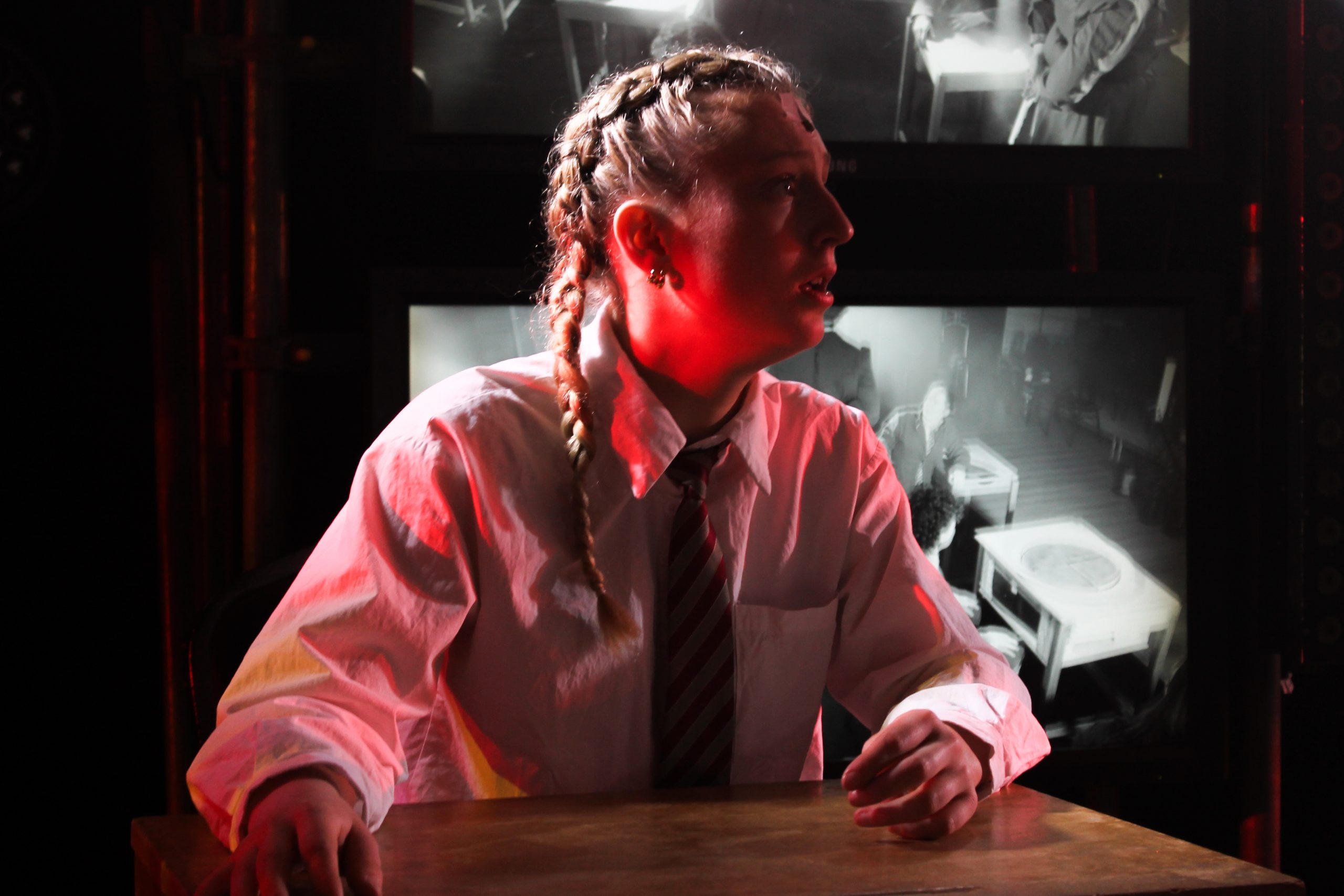 A girl with plaits in school uniform sits at a desk lit in red, looking up.