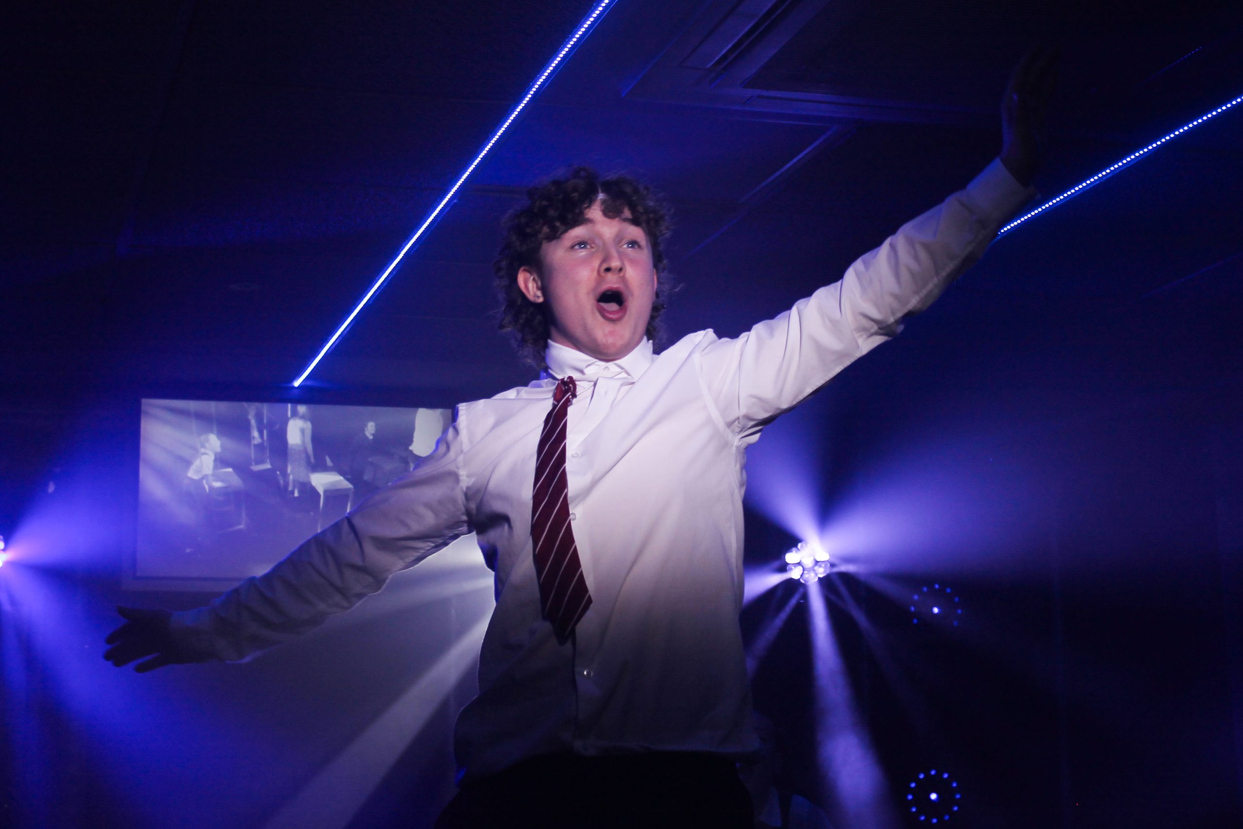 A boy in a school shirt and tie sings with arms outstretched as laser beams cross behind him.