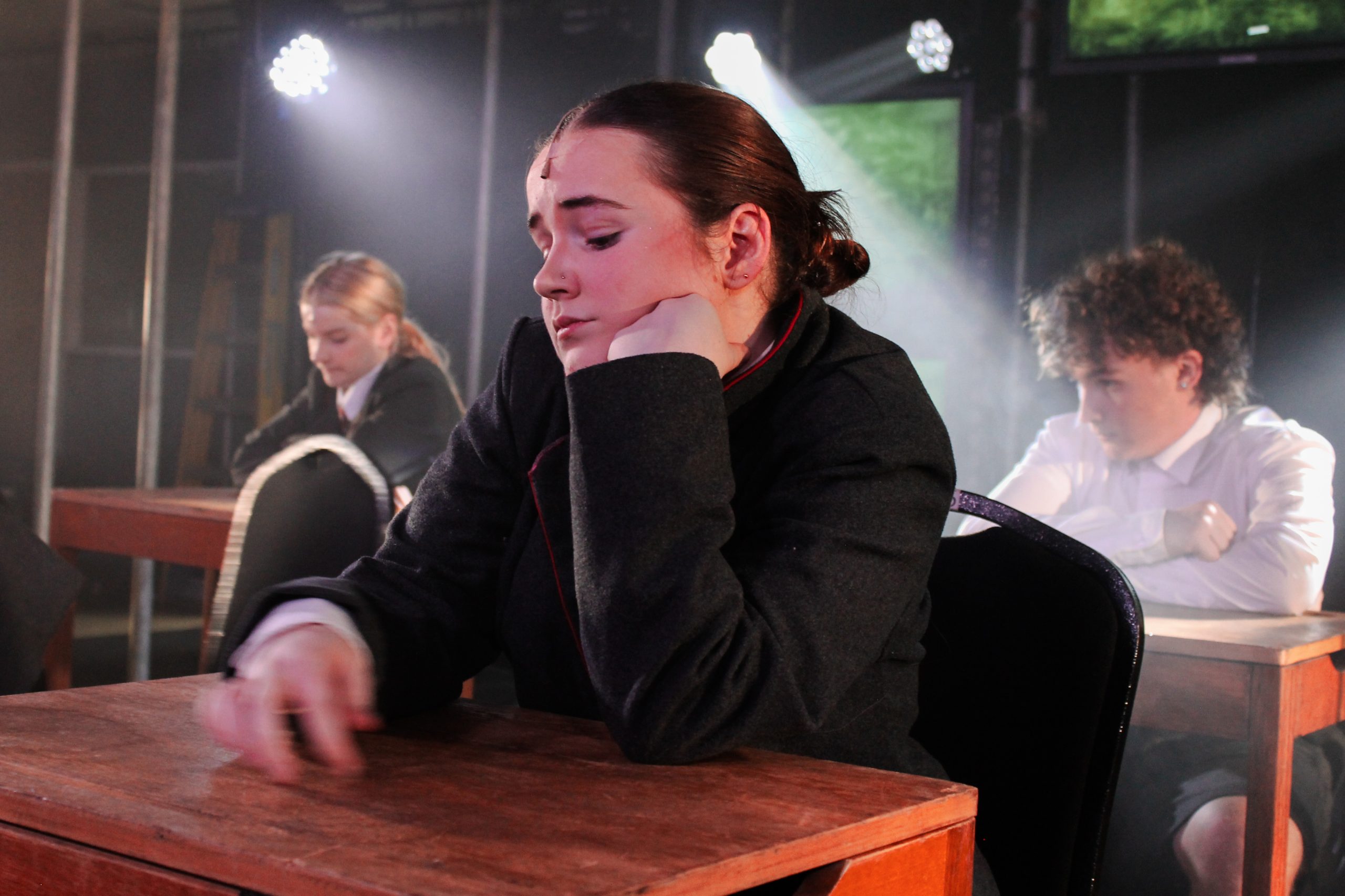 A pupil leans on her desk with her eyes closed while classmates sit behind her in a hazy classroom.