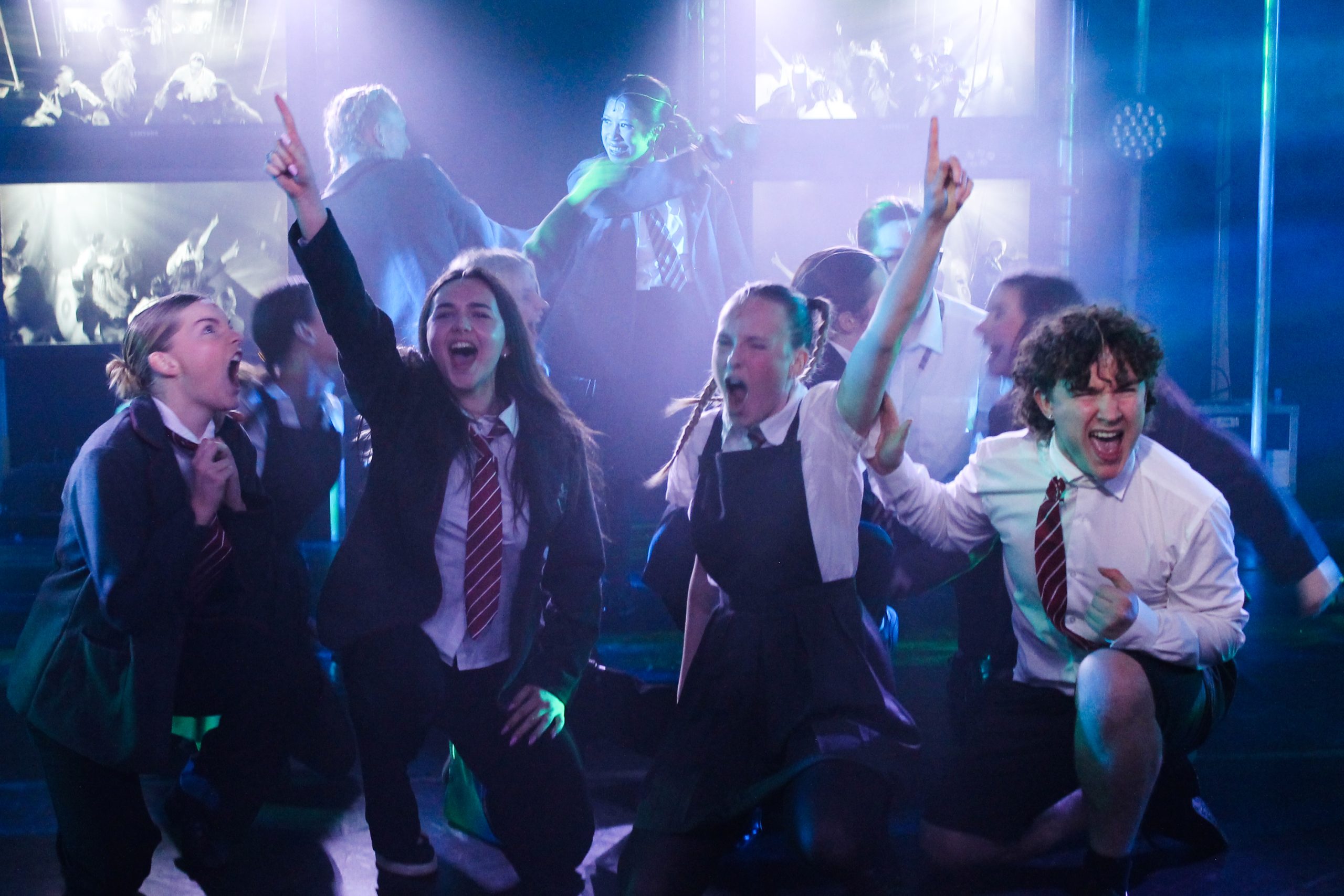 Pupils in school uniform cheer and point upwards in a lively ensemble number.