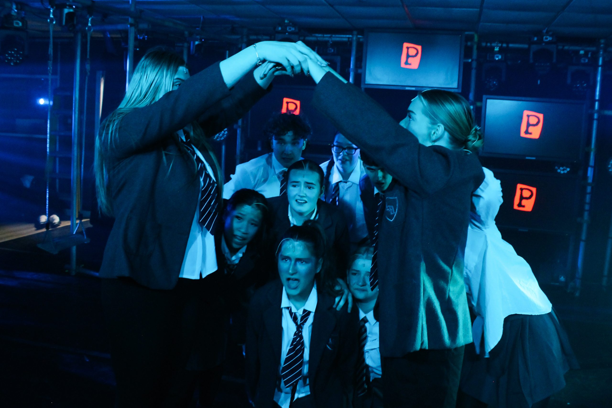 Pupils in school uniform huddle beneath raised arms, with letter blocks on screens behind.