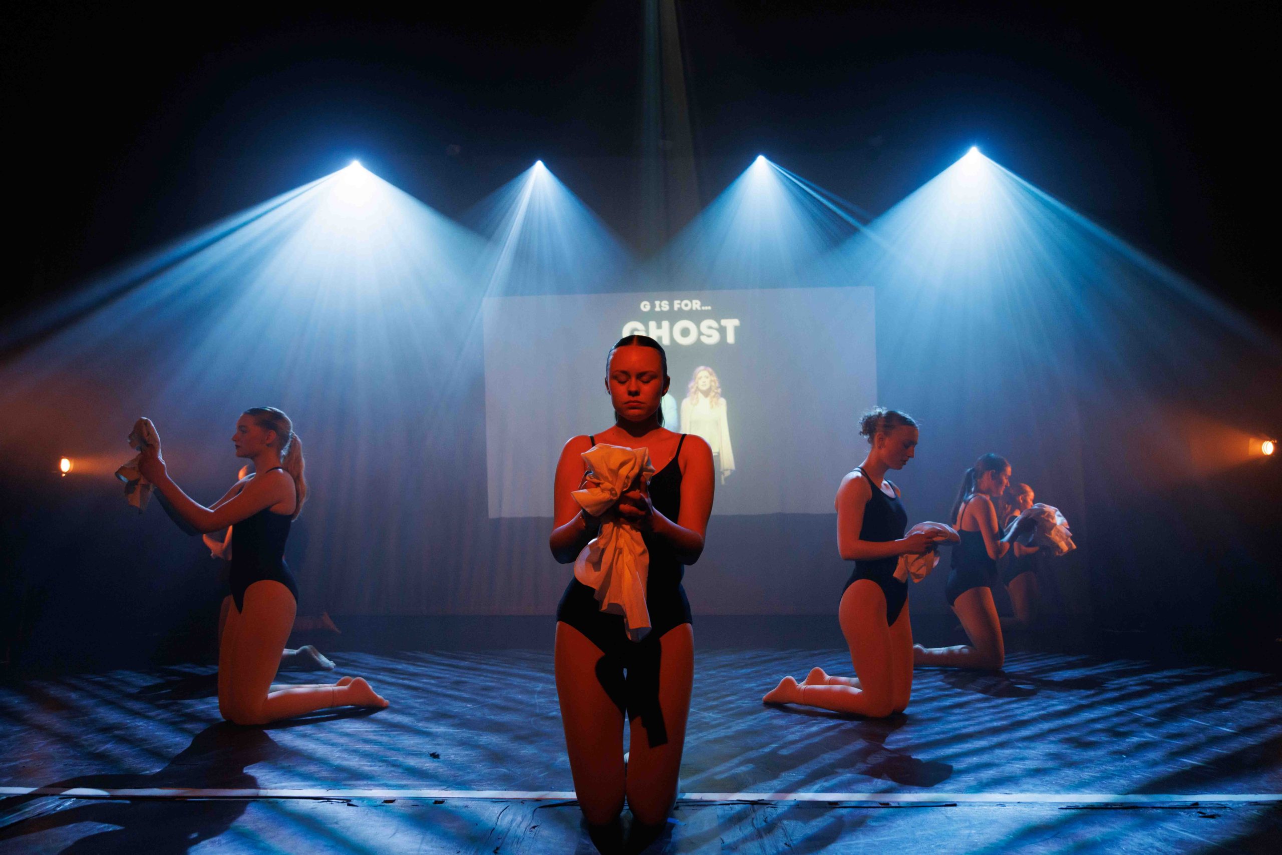Dancers kneel around a performer centre stage in front of a screen reading 'G is for Ghost'.