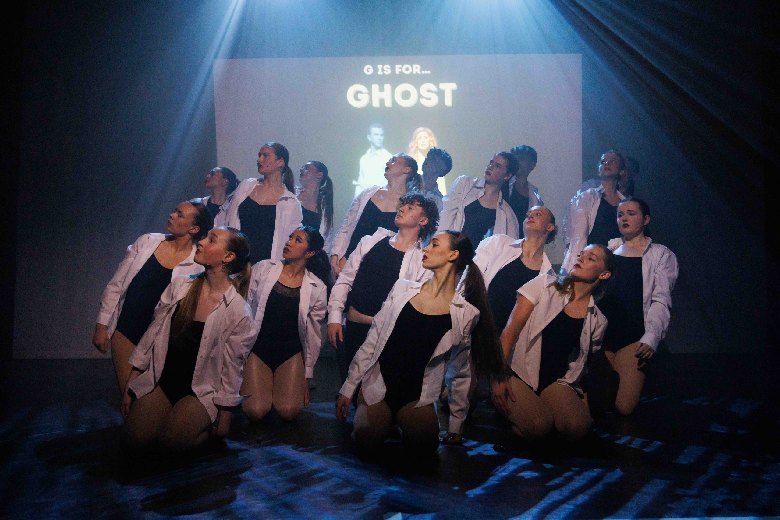 A large group of dancers in black leotards and white shirts kneel in formation in front of a 'Ghost' title screen.