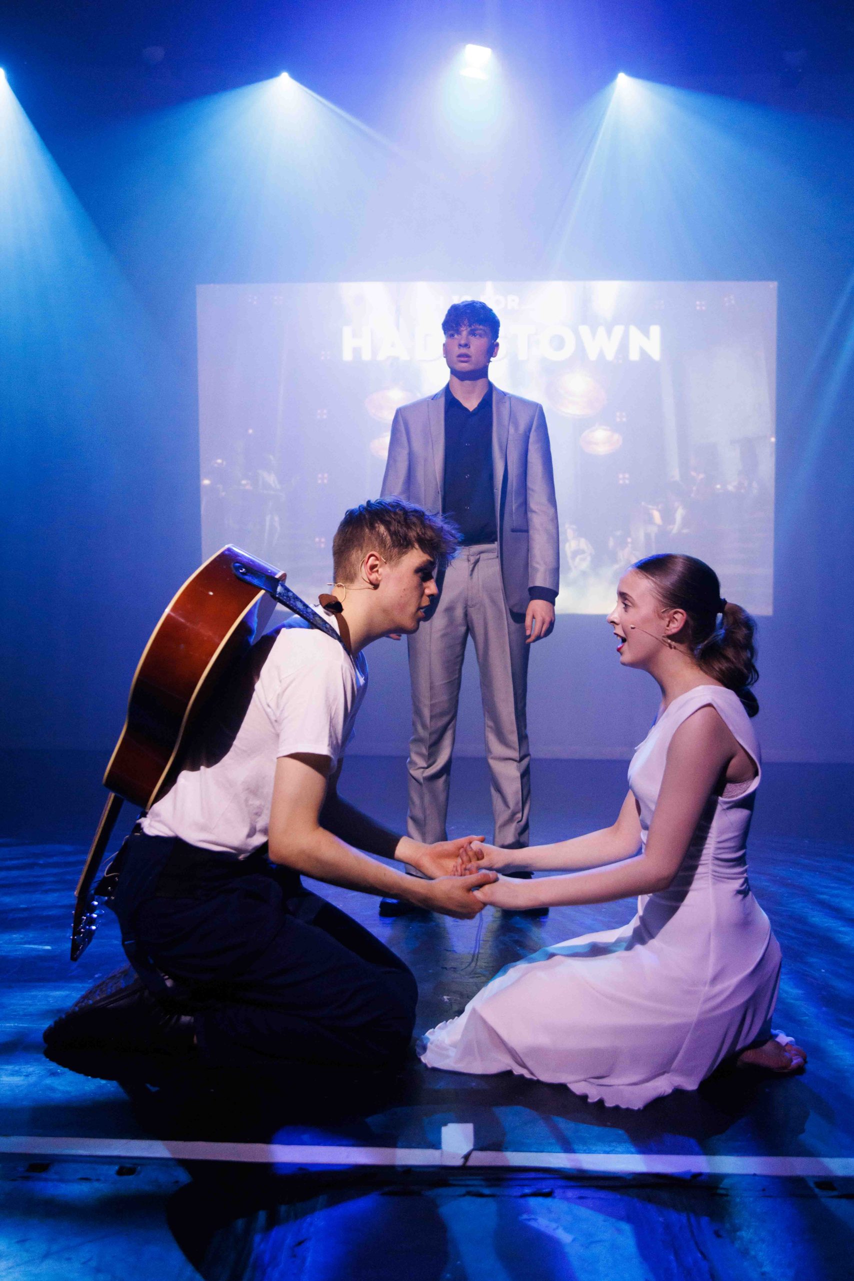 A young man with a guitar kneels holding hands with a young woman in white, as a man in a grey suit looks on, in front of a Hadestown title screen.