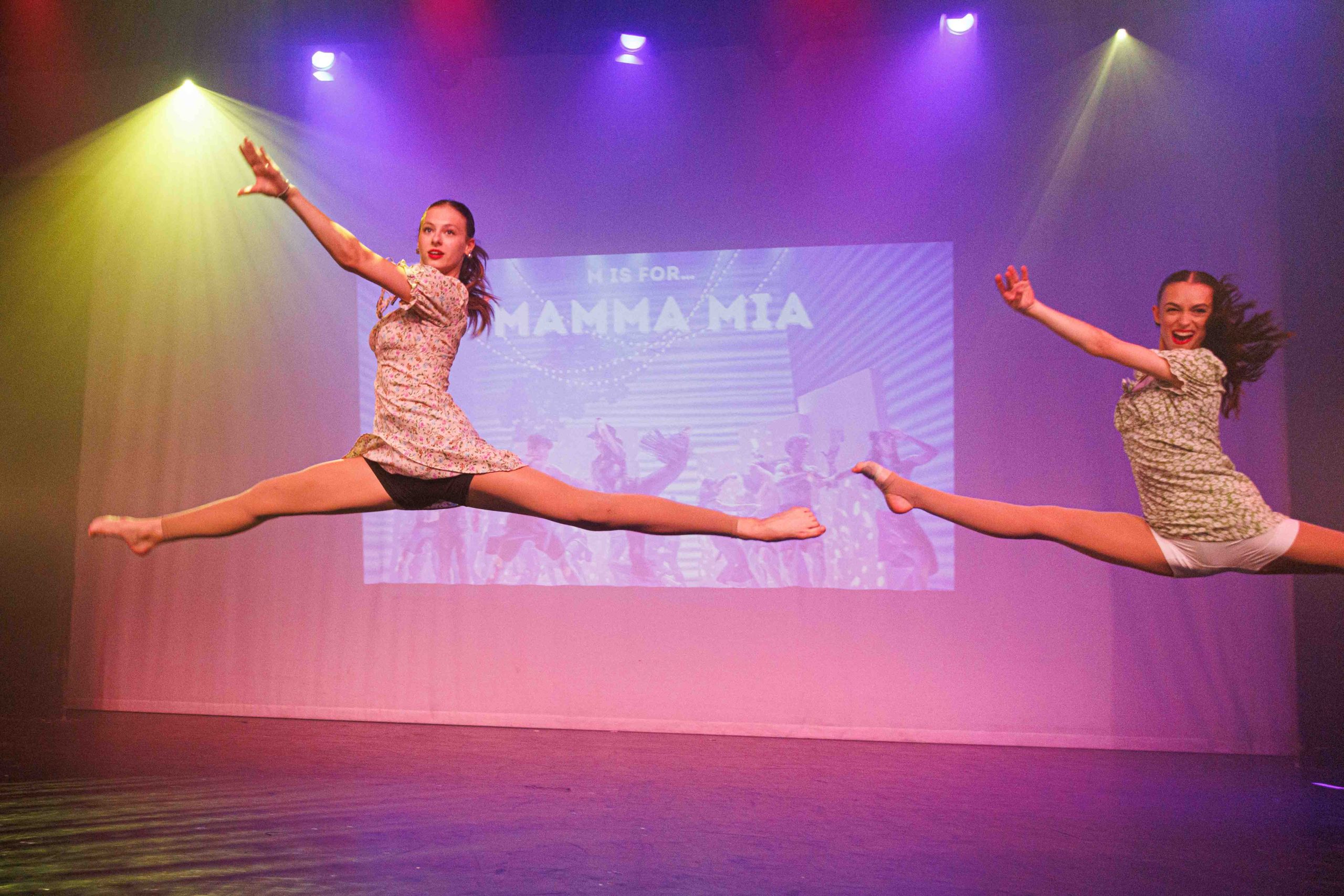 Two dancers in floral dresses leap into the splits in front of a Mamma Mia title screen.
