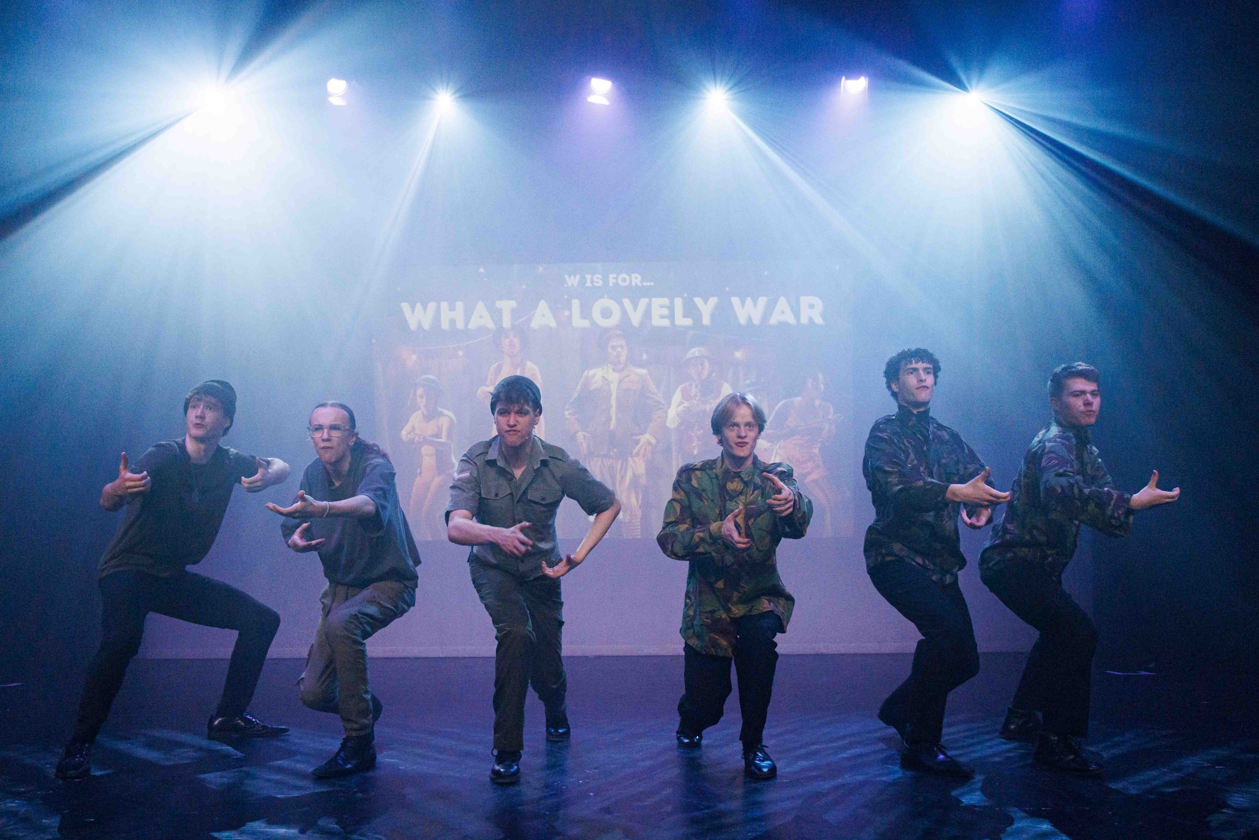 Performers in army camouflage crouch in a line in front of a screen reading 'W is for What a Lovely War'.
