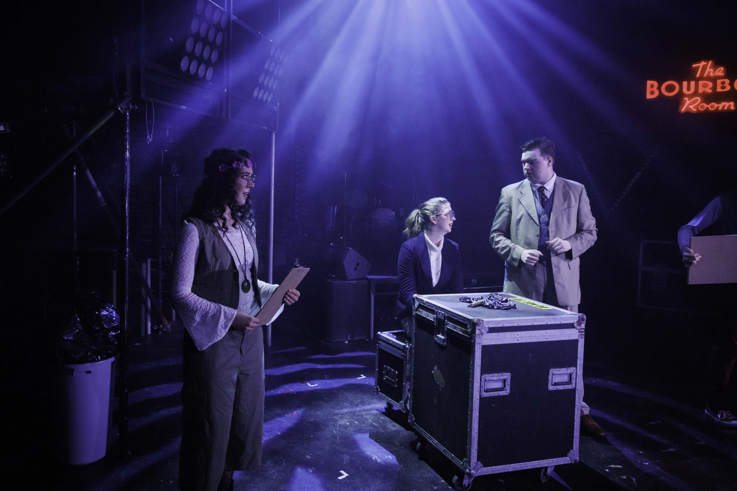 Three performers stand around a flight case beneath 'The Bourbon Room' neon sign.