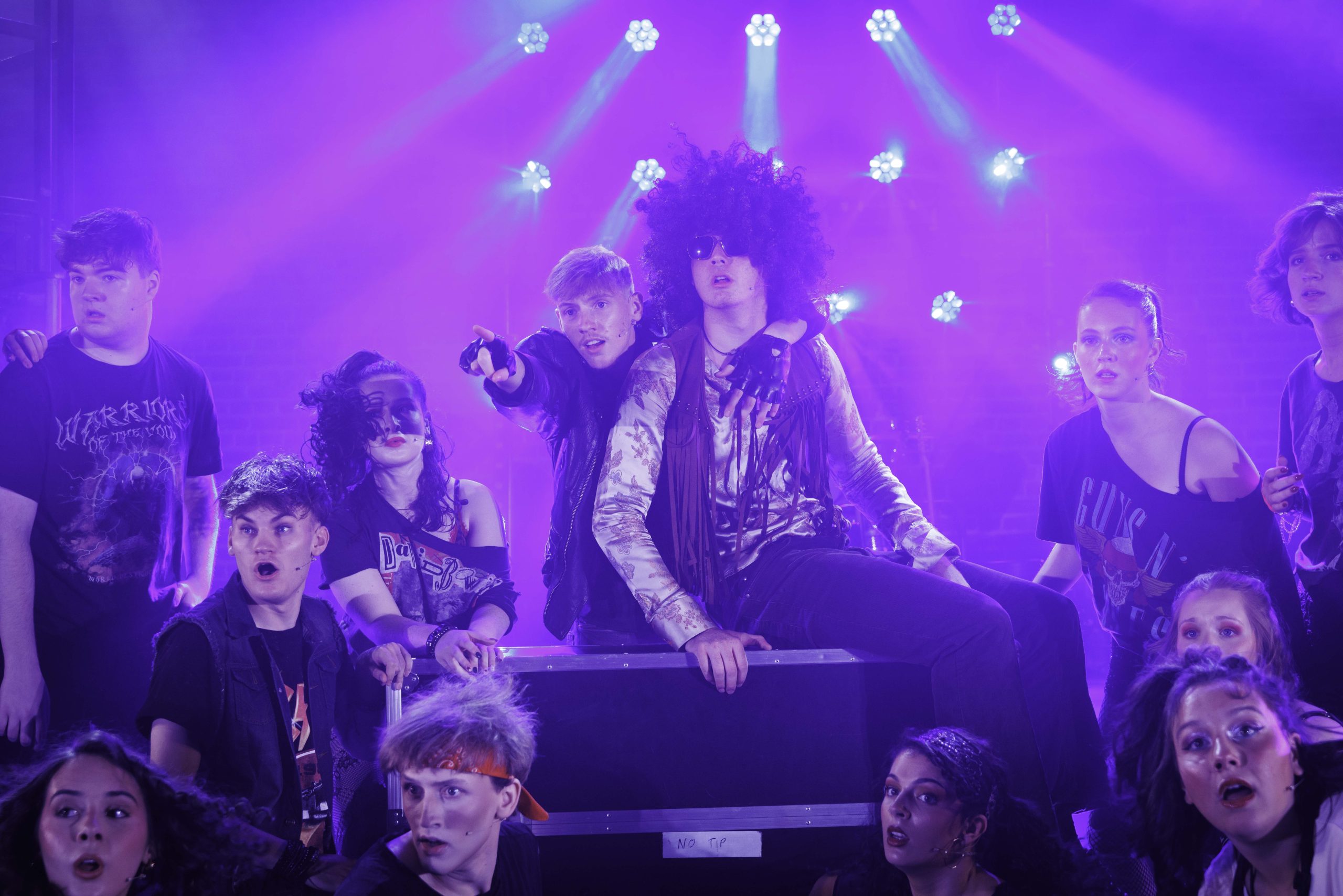 Rockers in wigs and band T-shirts gather around a performer in a big curly wig, pointing and reacting.