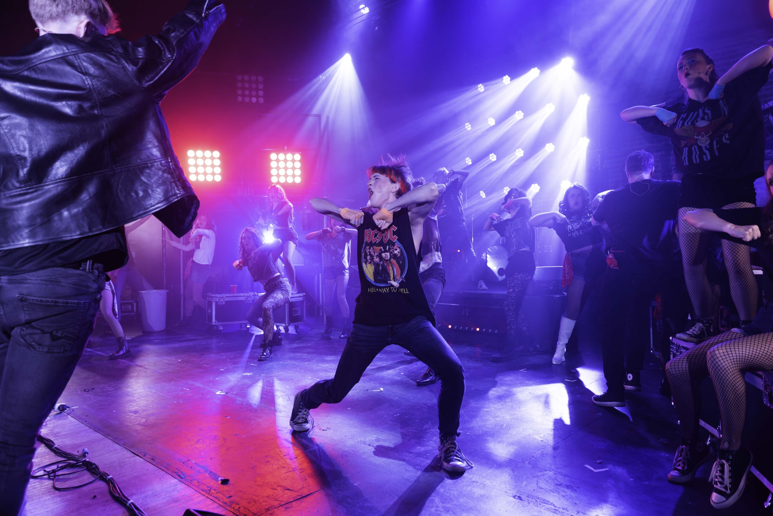 Performers in band T-shirts dance energetically on a red and blue lit club set.