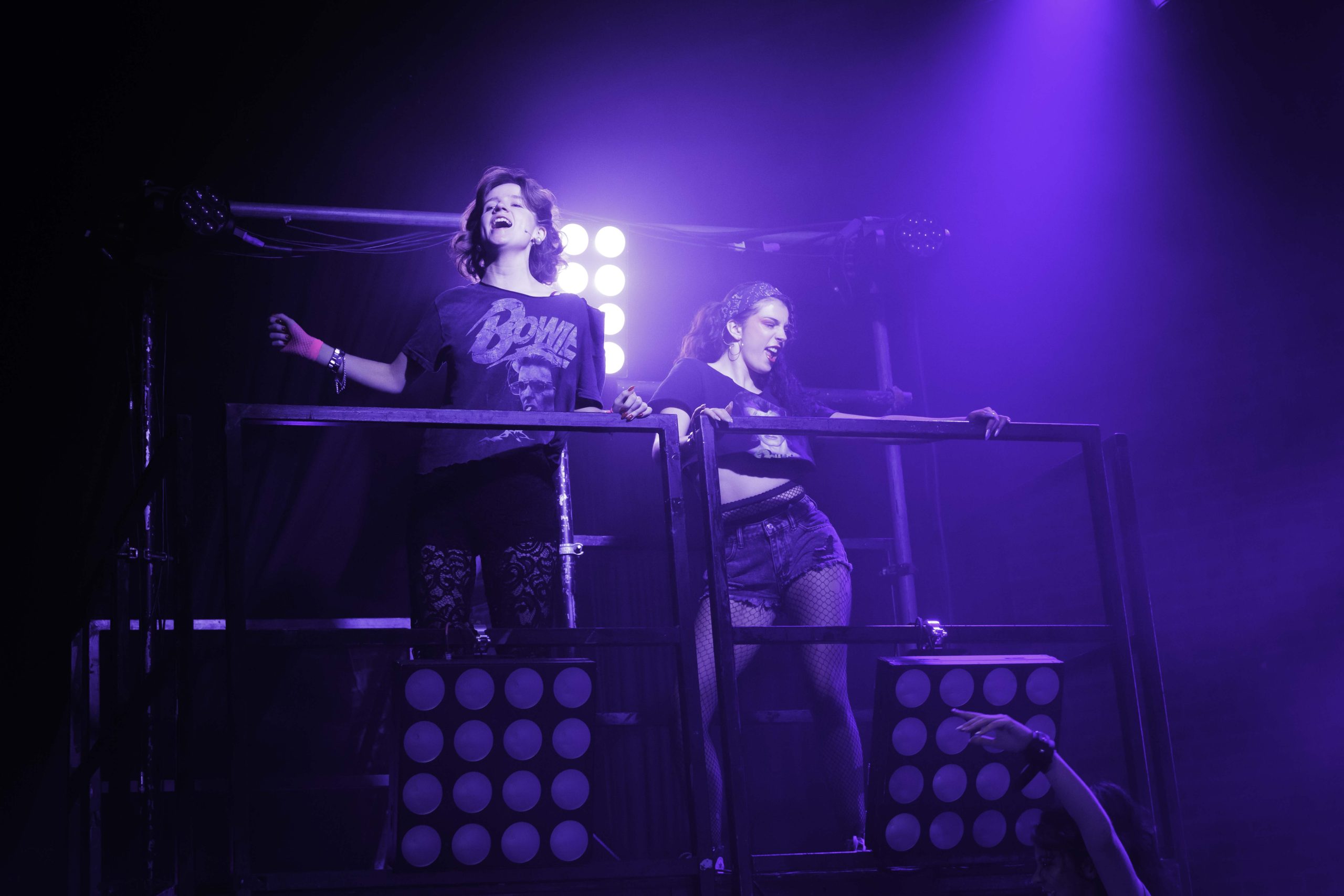 Two performers in rock T-shirts lean on a scaffold balcony above stage lights.