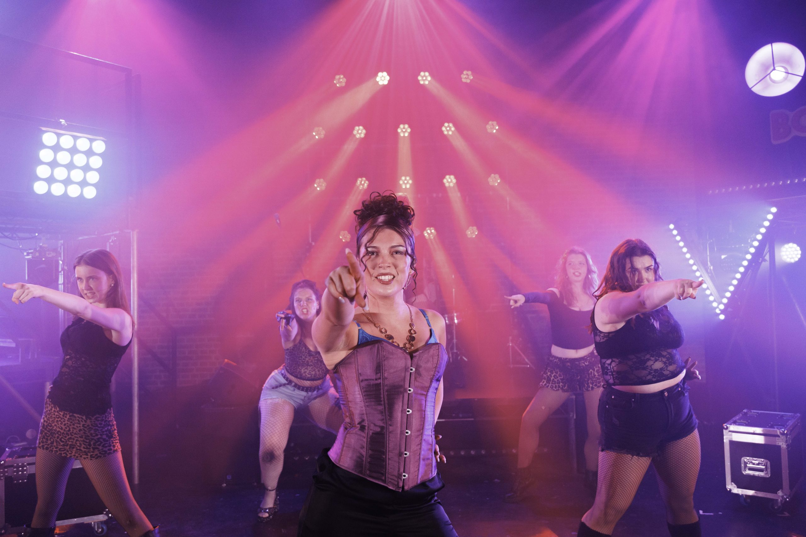 Performers in rock costumes point towards the audience under bright pink and red lights.
