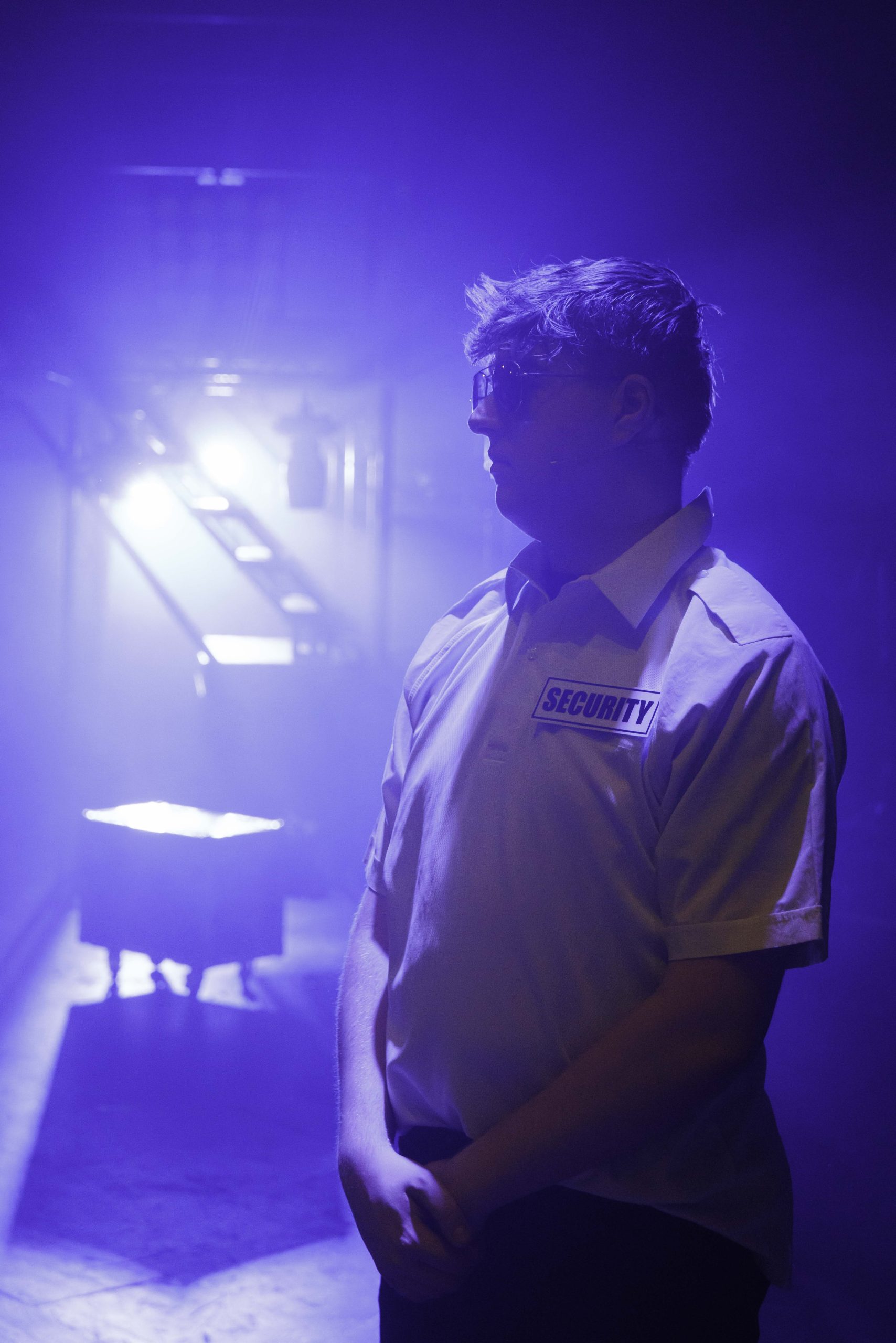 A performer in a security shirt stands in profile in blue backlight.