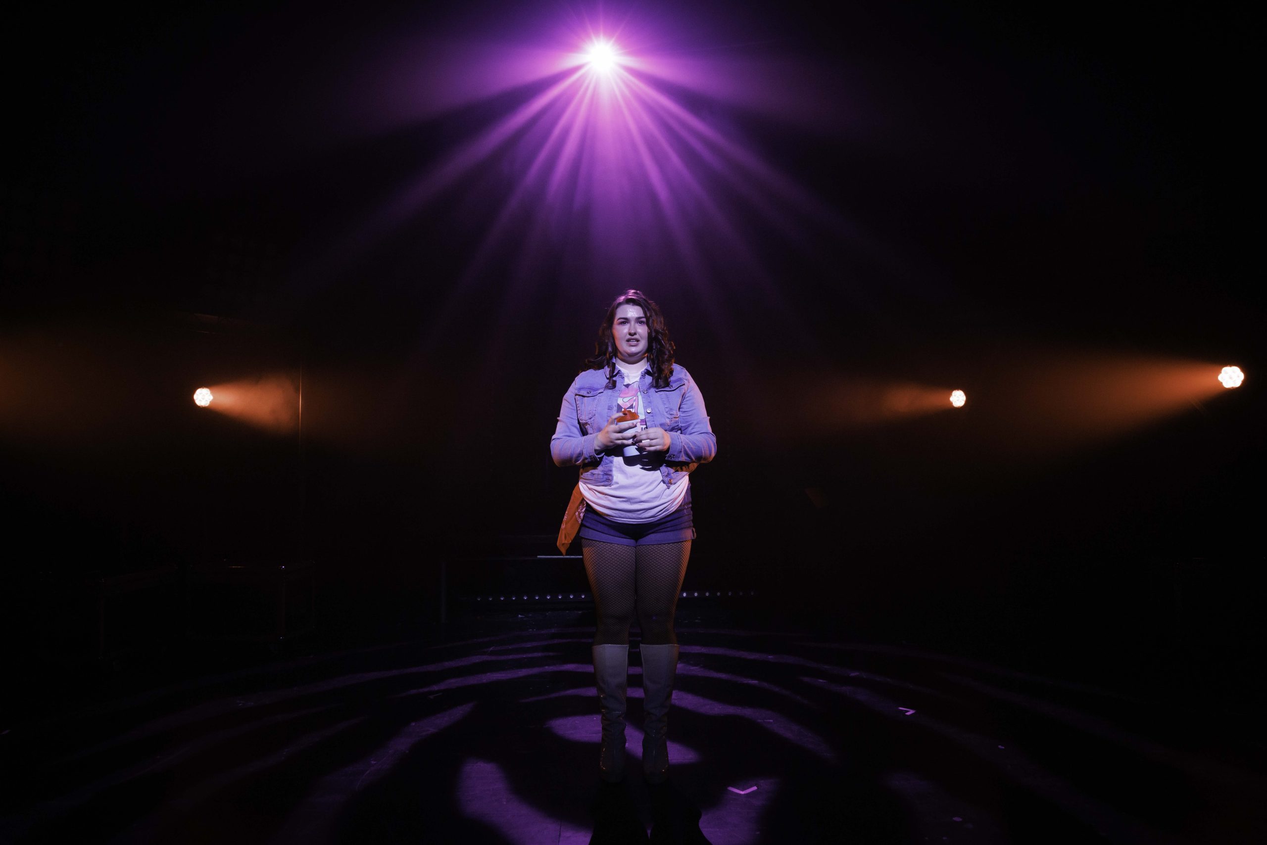 A performer in a grey hoodie stands alone in a pool of light on a dark stage.