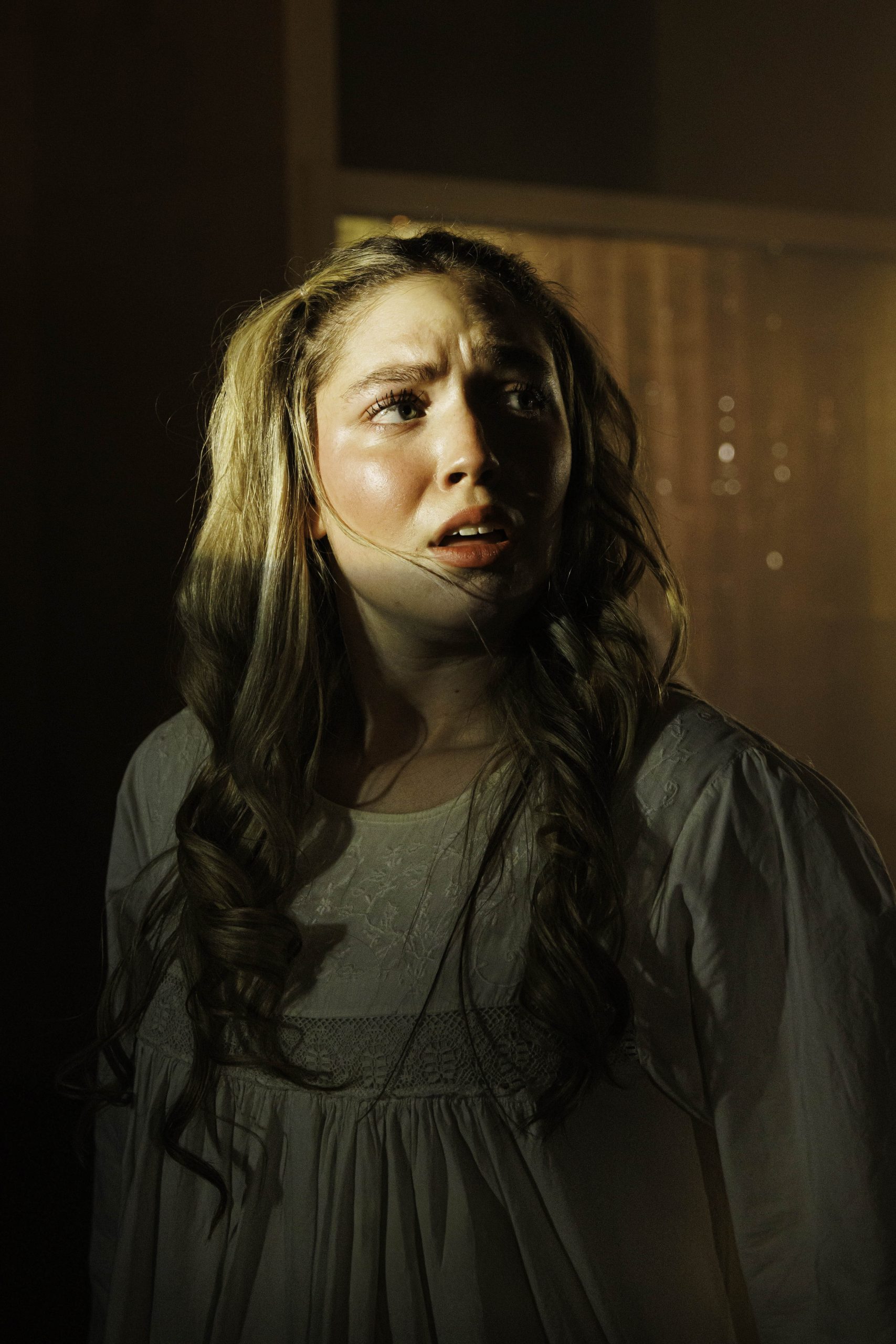 Close-up of a young woman with long curly hair looking upward in warm, low light.