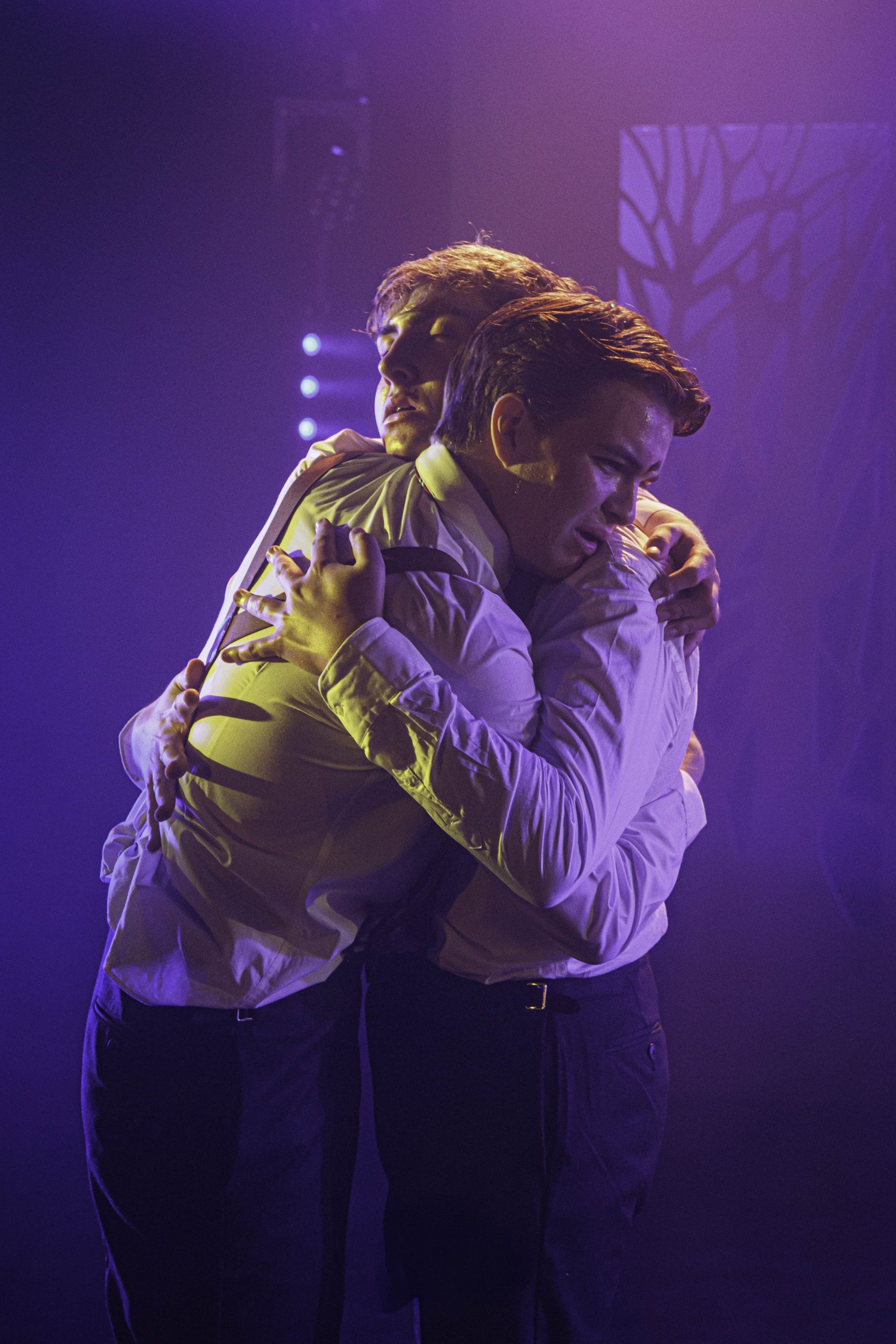 Two performers in white shirts embrace in purple light.