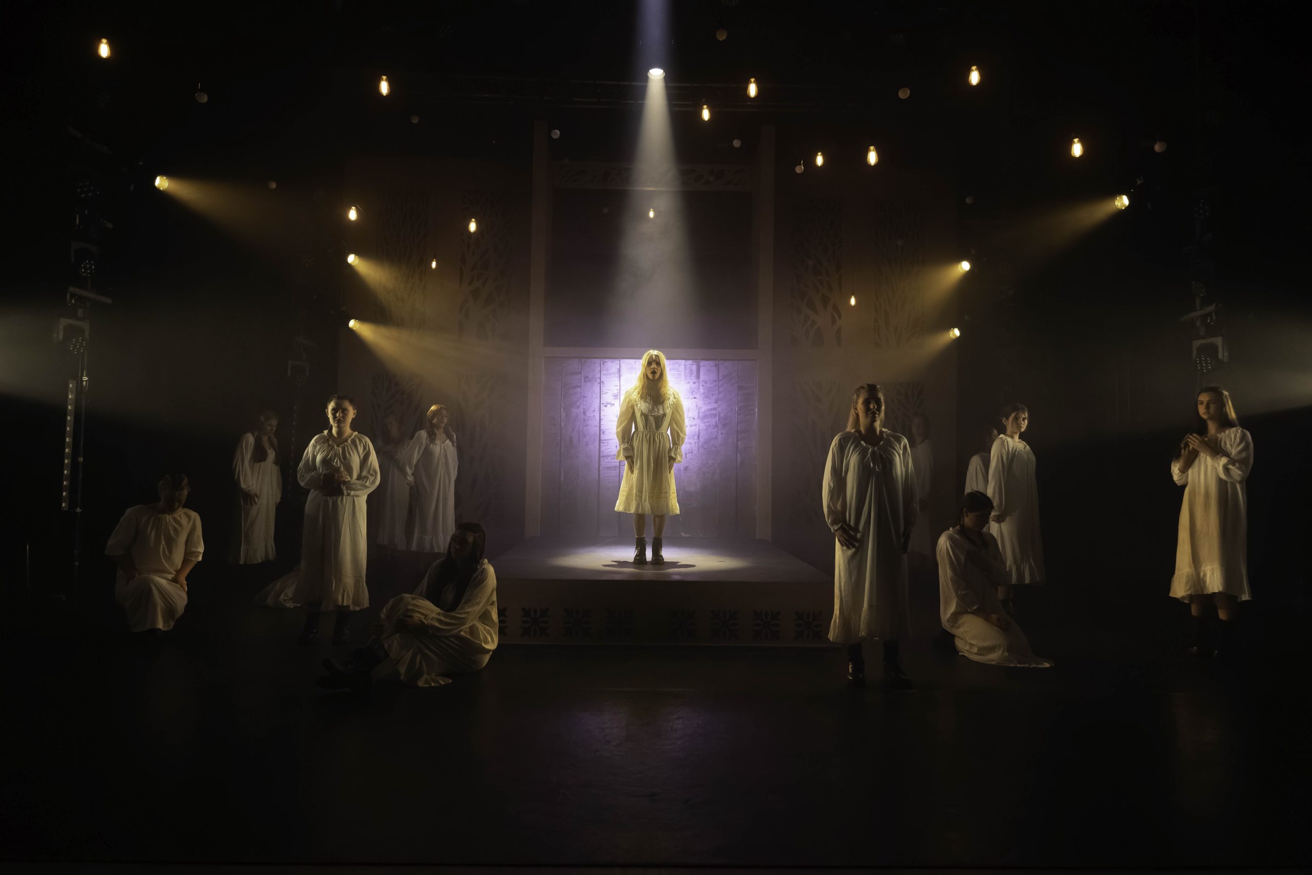 Performers in white nightgowns stand around a figure lit in a glowing doorway on a dark stage.