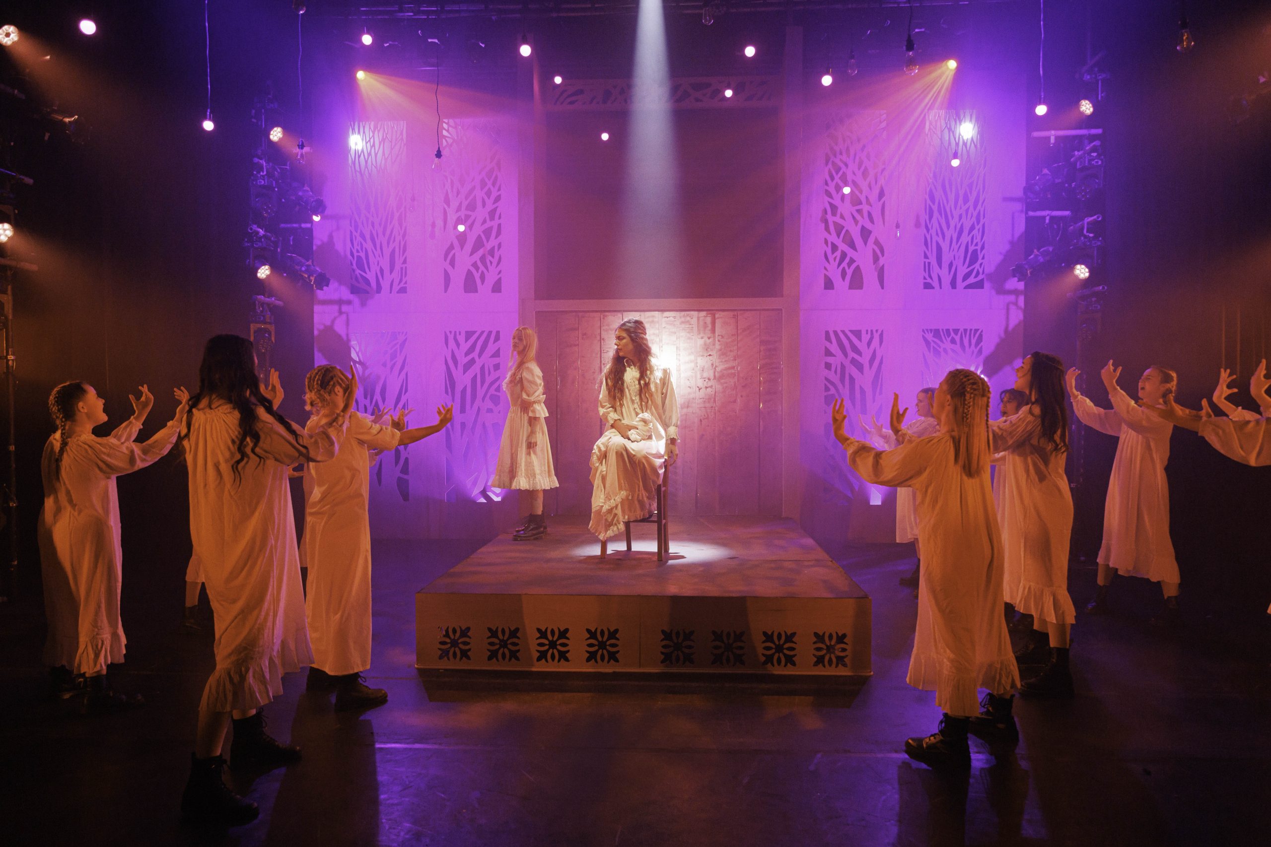 Performers in cream nightgowns reach towards a girl seated on a raised platform in purple light.