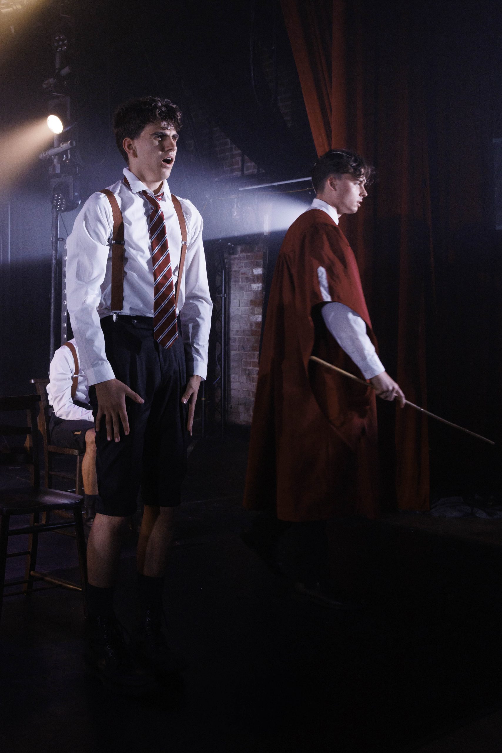 A schoolboy in shirt, tie and braces stands rigidly beside a teacher in a red coat holding a cane.