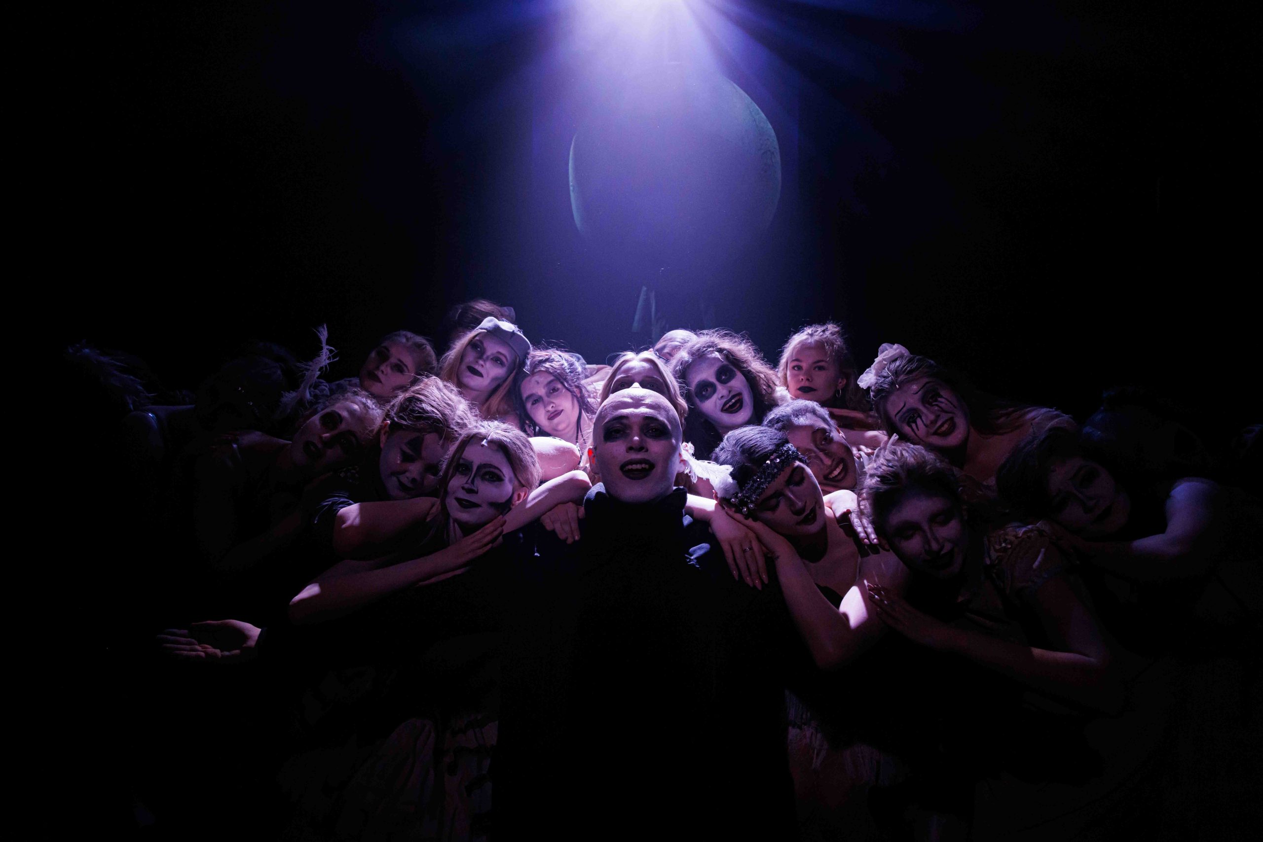 The Ancestors, in grey make-up, huddle tightly together under a single spotlight on a dark stage.