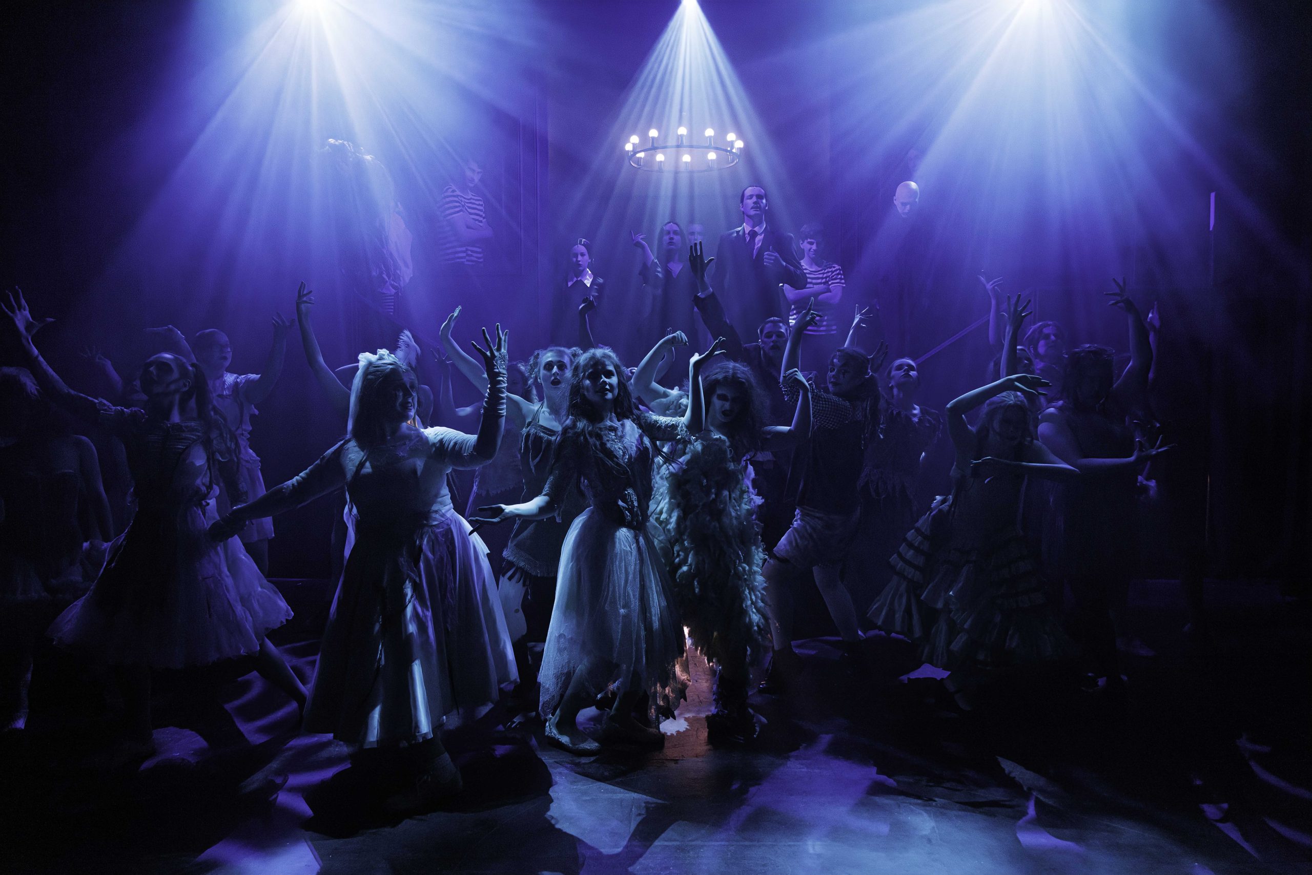 The Ancestors raise their arms in a ghostly tableau under blue spotlights and a glowing chandelier.