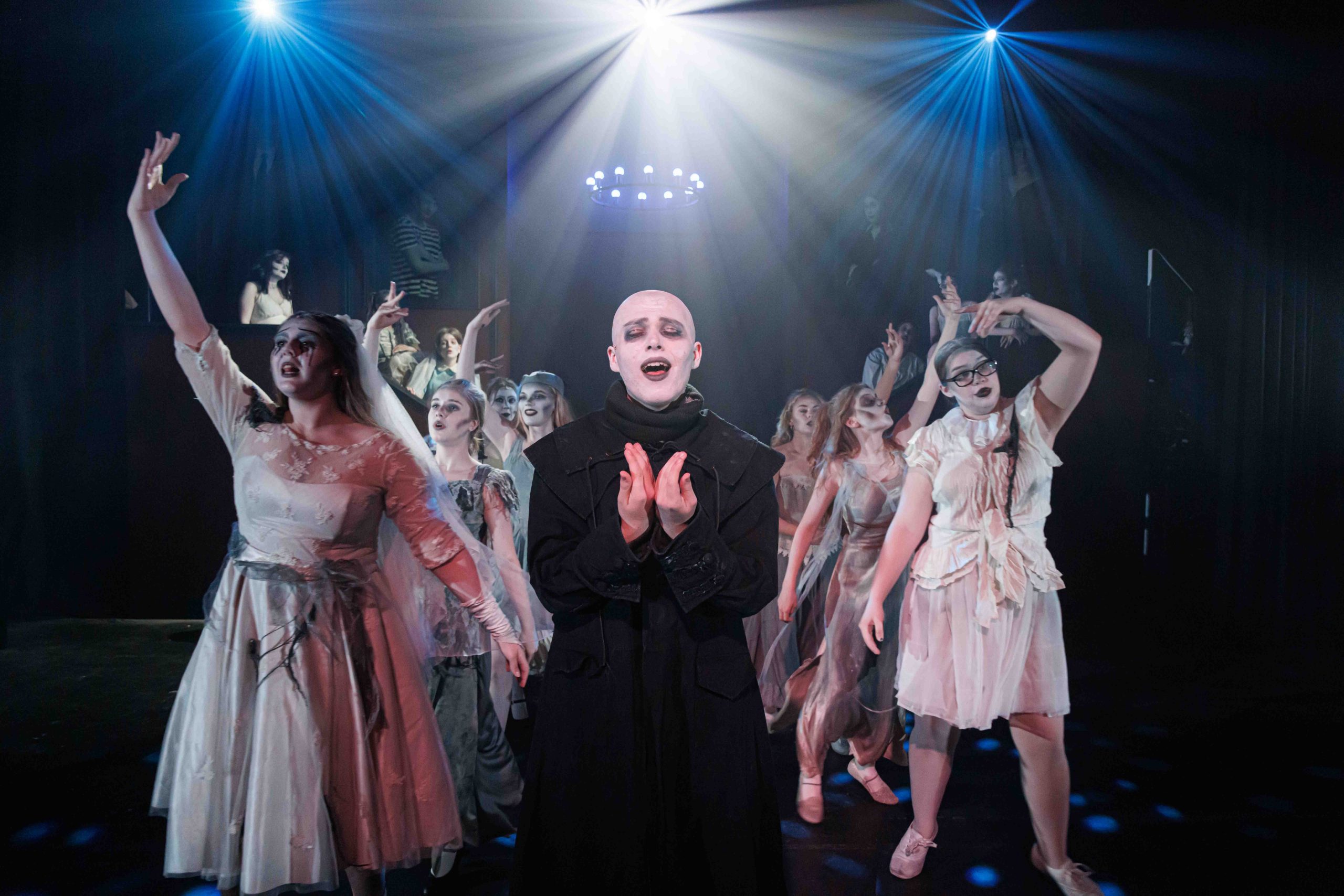 Uncle Fester, pale and bald in a black robe, sings with hands clasped, surrounded by Ancestors in ghostly white costumes.