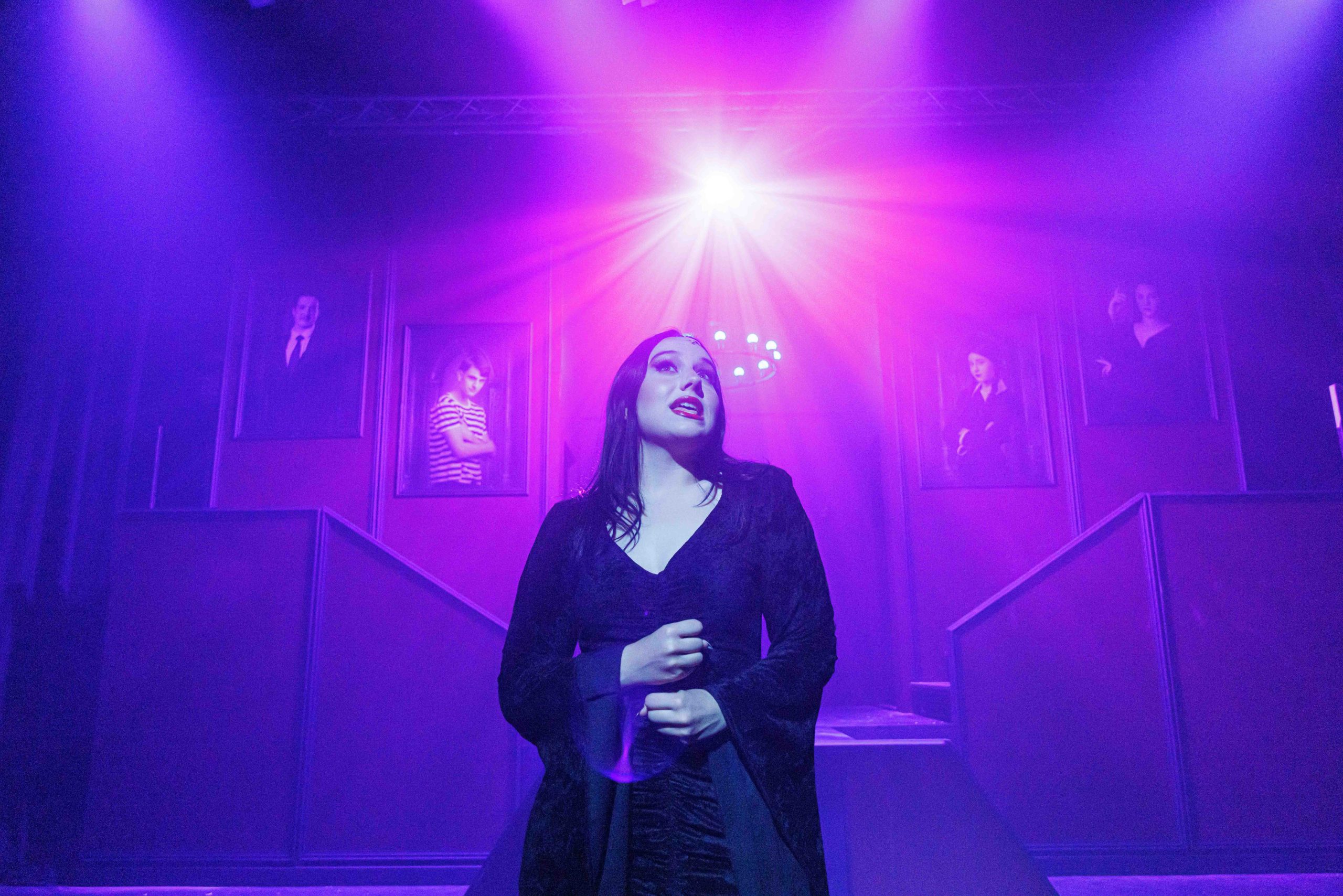 Morticia, in a long black gown, stands centre stage under a bright purple spotlight in front of the mansion staircase.