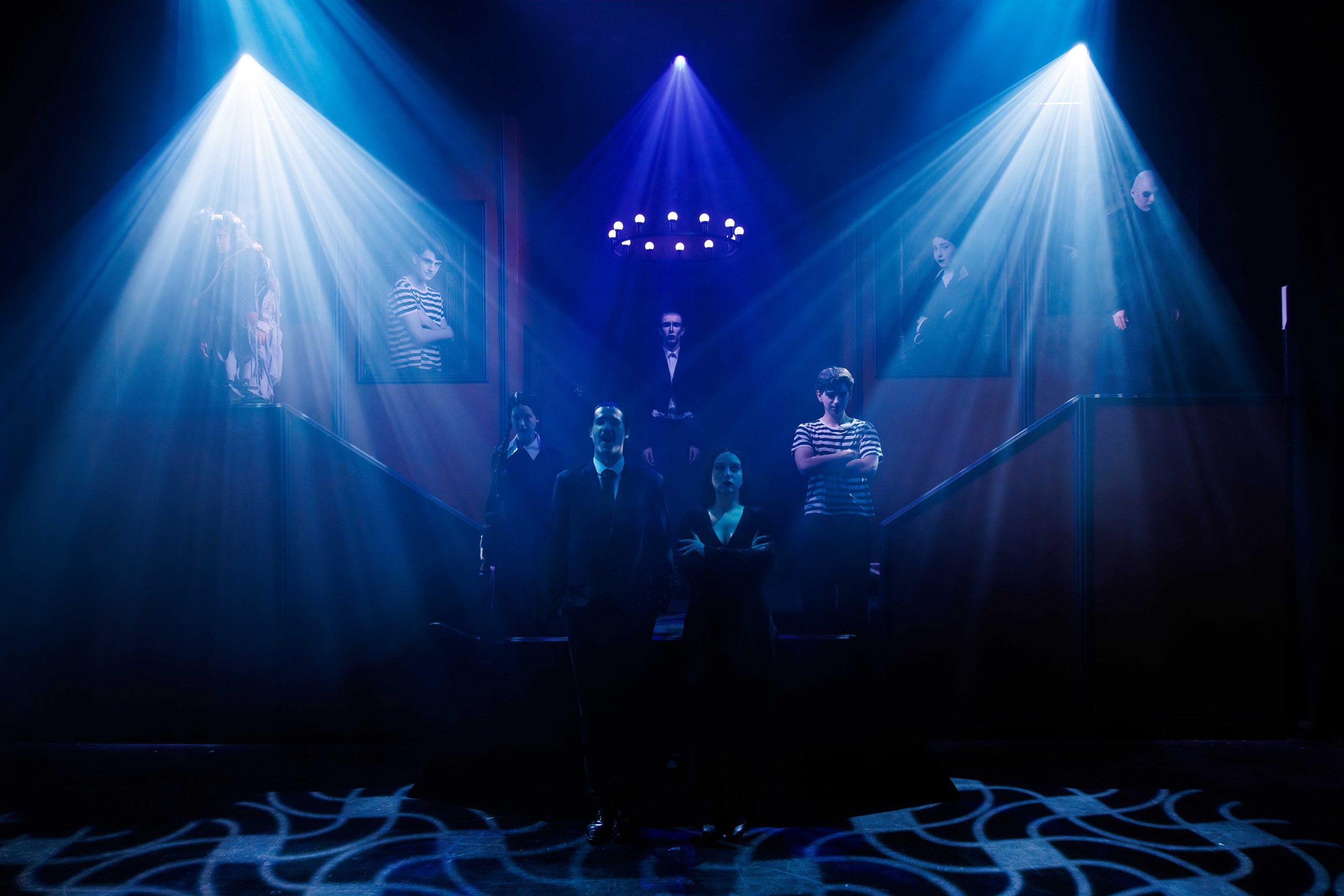 The Addams family stand in silhouette at the foot of the mansion staircase, lit by blue beams beneath a chandelier.