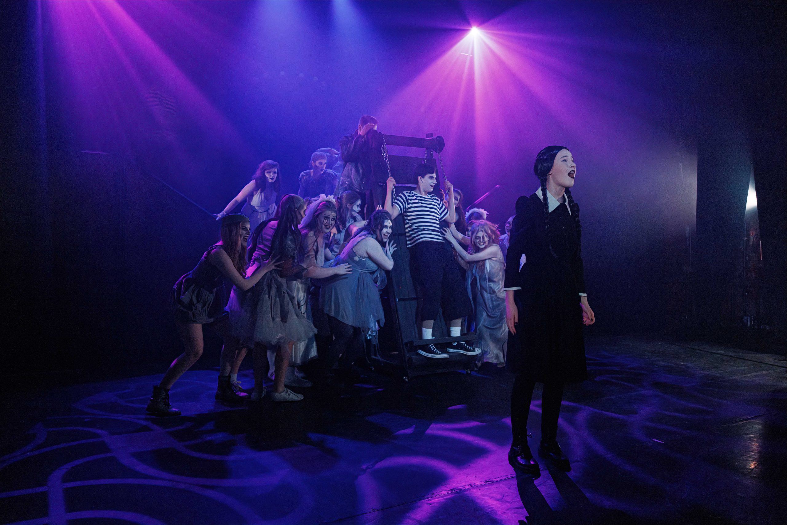 Wednesday, in a black dress, stands at the front of the stage as the ensemble lunges towards Pugsley behind her.